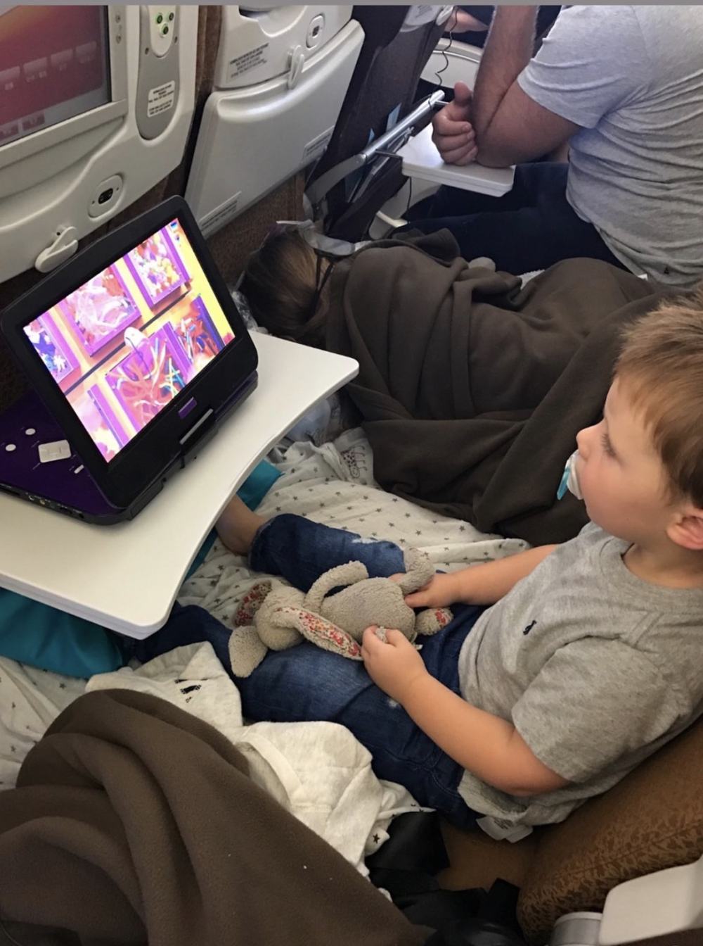 Jack sitting on a Kooshion in an economy seat, watching a screen, with his sister asleep beside him